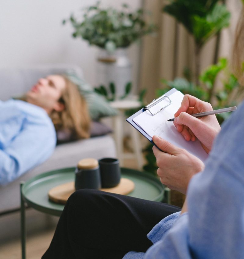 A therapy session in progress with a therapist taking notes as a patient reclines on a sofa.